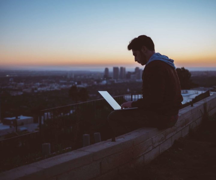 
				“A young man working on a laptop while seated on a ledge in a city during sunset” by Avi Richards on Unsplash		