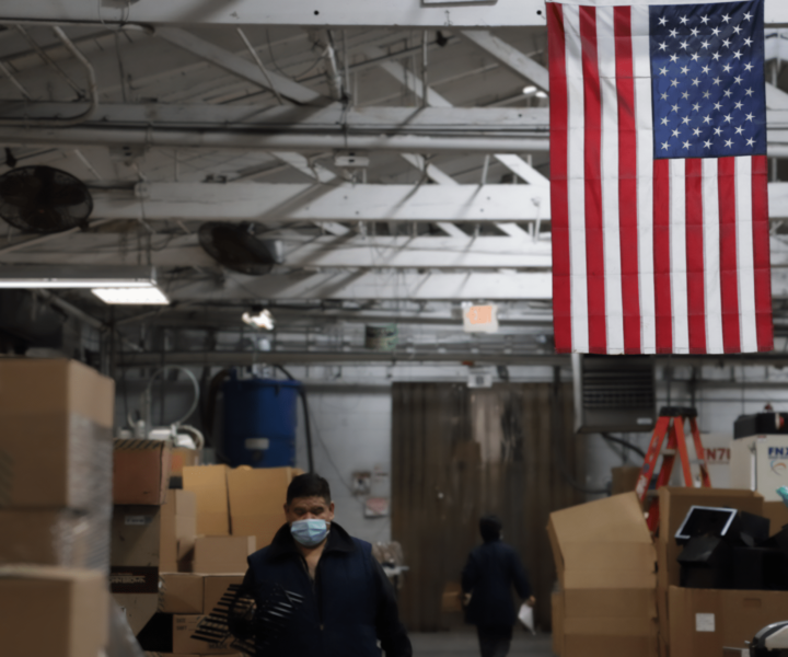 A man walks toward the camera through a warehouse and passes under an American Flag hanging from the rafters of the plastic comb factory. He is wearing a blur surgical mask as a woman walks away from him and the camera. Photo by Julian Rigg.