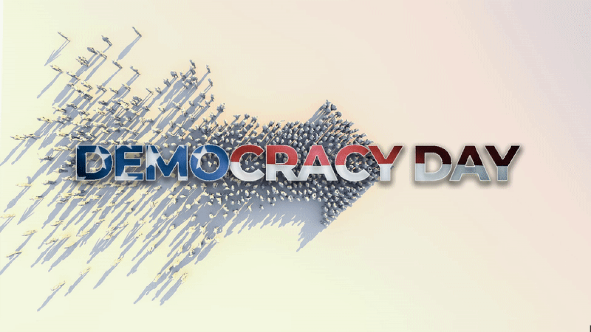 DECORATION ONLY: A birds-eye view of a crowd of people standing in the shape of an arrow pointing to the right with the words DEMOCRACY DAY over their heads, filled with a waving American flag.