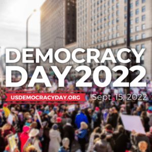 A crowd of demonstrators gathers in the streets in the background of the image, with the Democracy Day 2022 logo in the foreground.