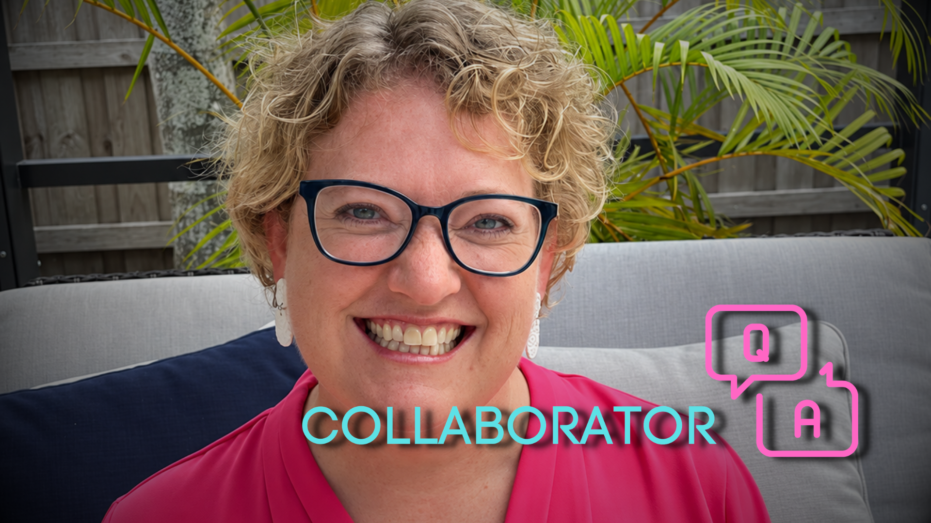Joy Mayer smiling with short curly blond hair and dark-framed glasses sits on a light gray outdoor sofa, wearing a bright pink blouse and white earrings, with green palm fronds and a wooden fence behind her.